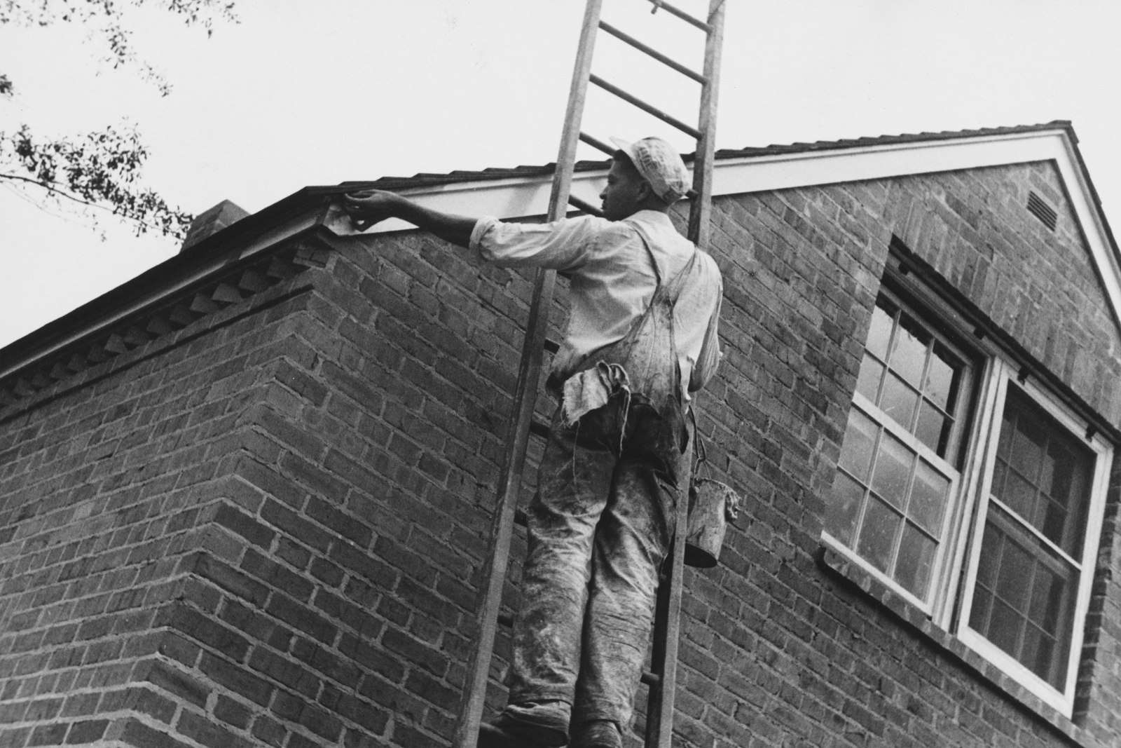 Painter on a tall ladder painting the eaves of a two-storey brick house, in an old black-and-white photo