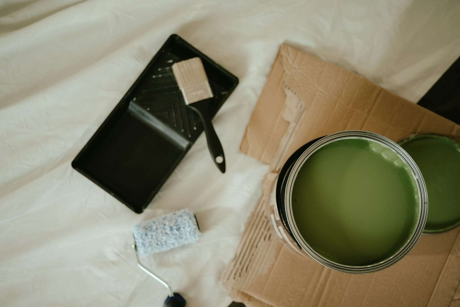Open tin of green paint with a roller, tray and brush on a drop sheet